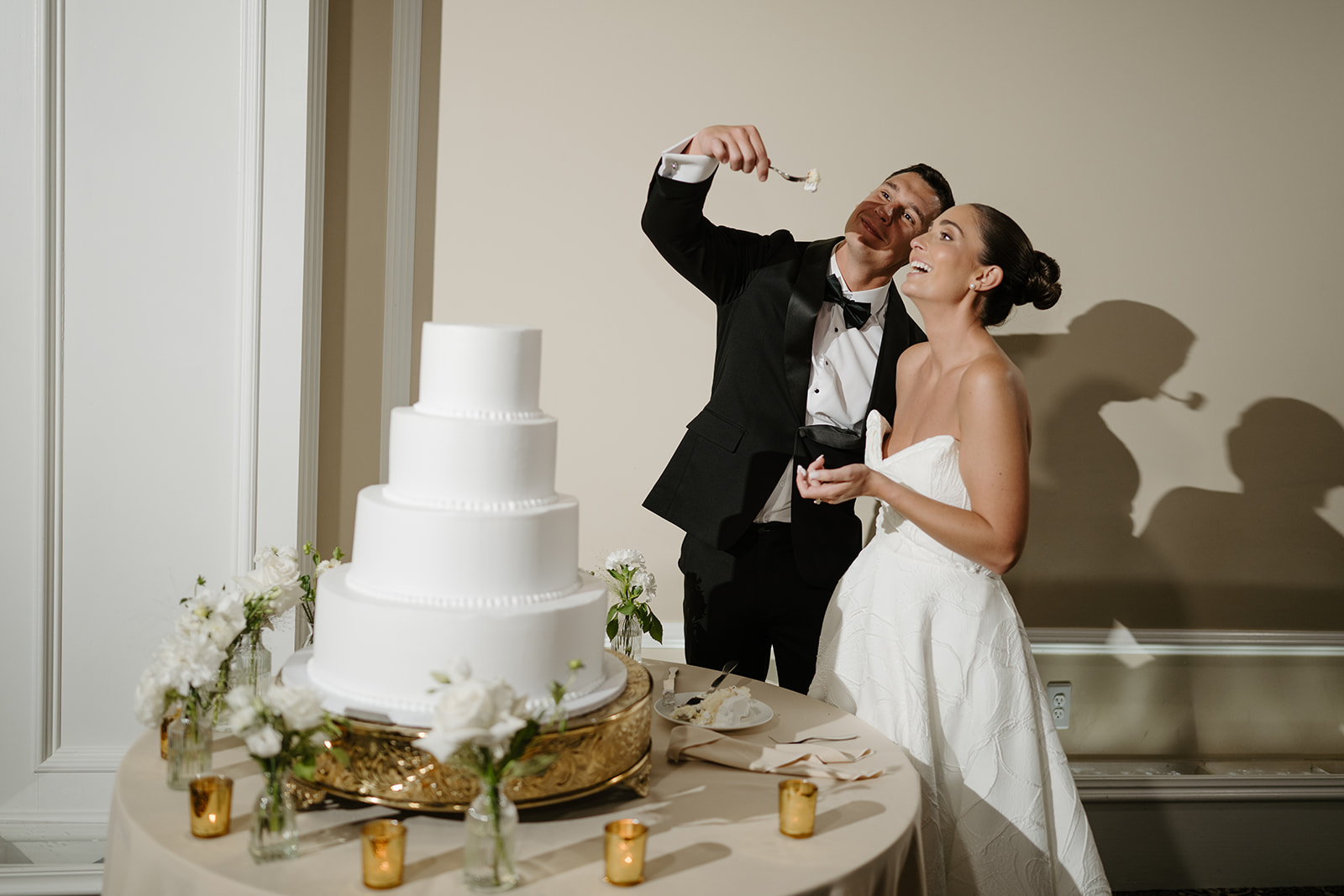 Groom playfully holding a bite of cake on a fork for the bride at their reception cake table.